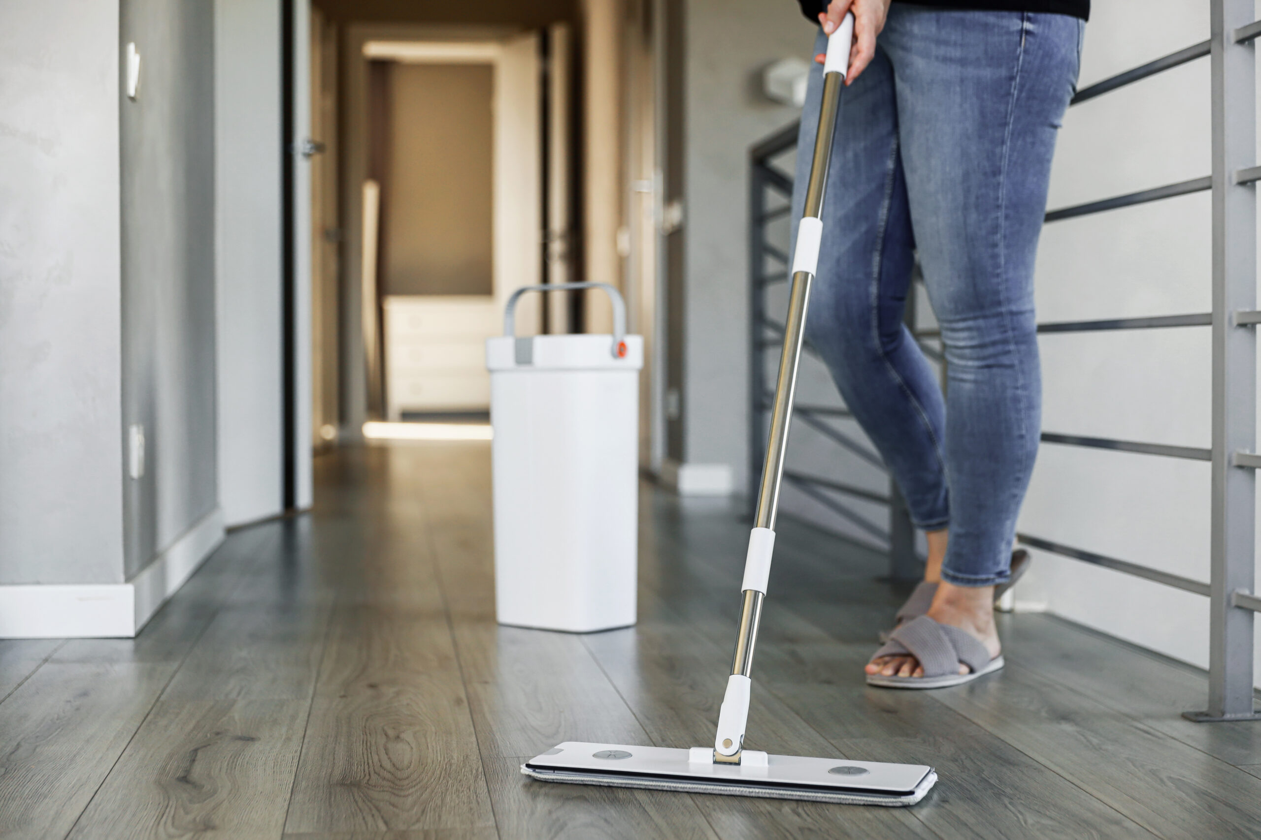 Pessoa com jeans e chinelos utilizando um esfregão moderno para limpar um piso de madeira ou vinílico em um corredor residencial, com um balde de limpeza branco ao lado.