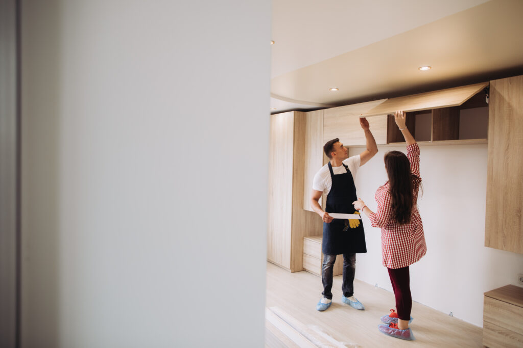 Casal inspecionando a instalação de armários embutidos de madeira em um cômodo, com o homem de avental segurando pranchetas e a mulher apontando para as portas superiores do móvel.