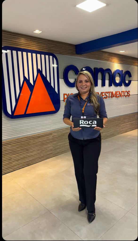 Colaboradora da Comac Materiais de Construção sorrindo e segurando uma placa da marca parceira Roca. Ela veste o uniforme da empresa (camisa social azul com logo) e está posicionada em frente ao painel institucional com o logotipo da Comac em destaque no fundo