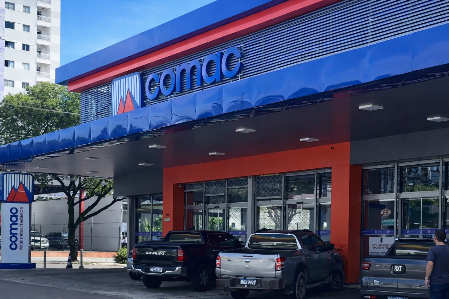 Fachada moderna da loja Comac Materiais de Construção em Manaus. A imagem destaca a estrutura metálica em azul e laranja, o logotipo da marca no topo e o estacionamento frontal com caminhonetes, representando um ponto de venda robusto e acessível.